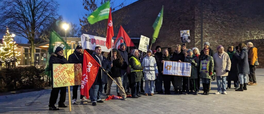 „Gruppe von Demonstrierenden mit Fahnen und Schildern auf einem Platz am frühen Abend; sie halten Banner politischer und zivilgesellschaftlicher Gruppen.“
