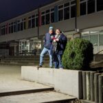 Zwei Personen stehen am Abend auf einem niedrigen Betonpodest vor einem Schul- oder Verwaltungsgebäude und halten ein Megafon.