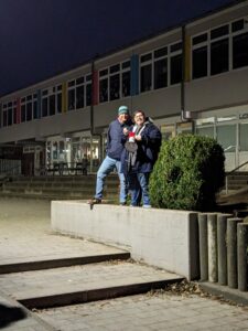 Zwei Personen stehen am Abend auf einem niedrigen Betonpodest vor einem Schul- oder Verwaltungsgebäude und halten ein Megafon.