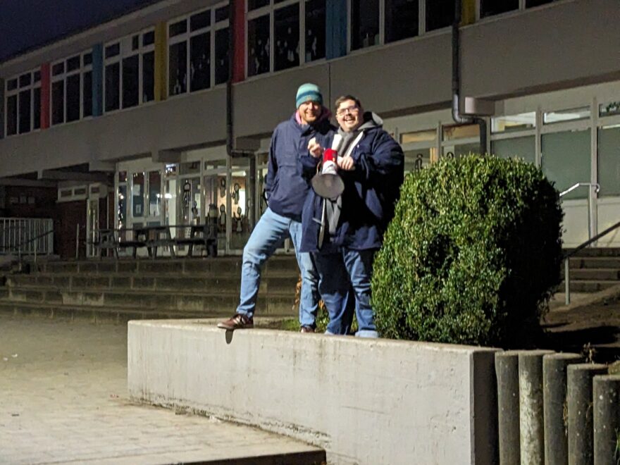 Zwei Personen stehen am Abend auf einem niedrigen Betonpodest vor einem Schul- oder Verwaltungsgebäude und halten ein Megafon.